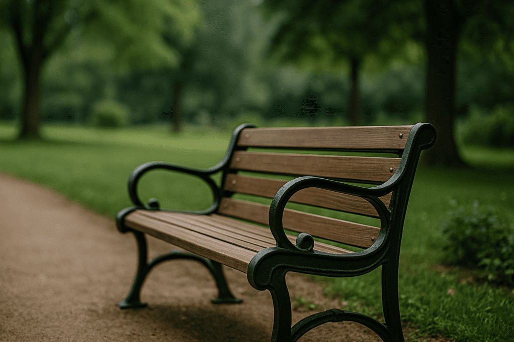 A wooden and metal park bench along a curving dirt path in a green, tree-lined park under overcast skies, with a blurred natural background