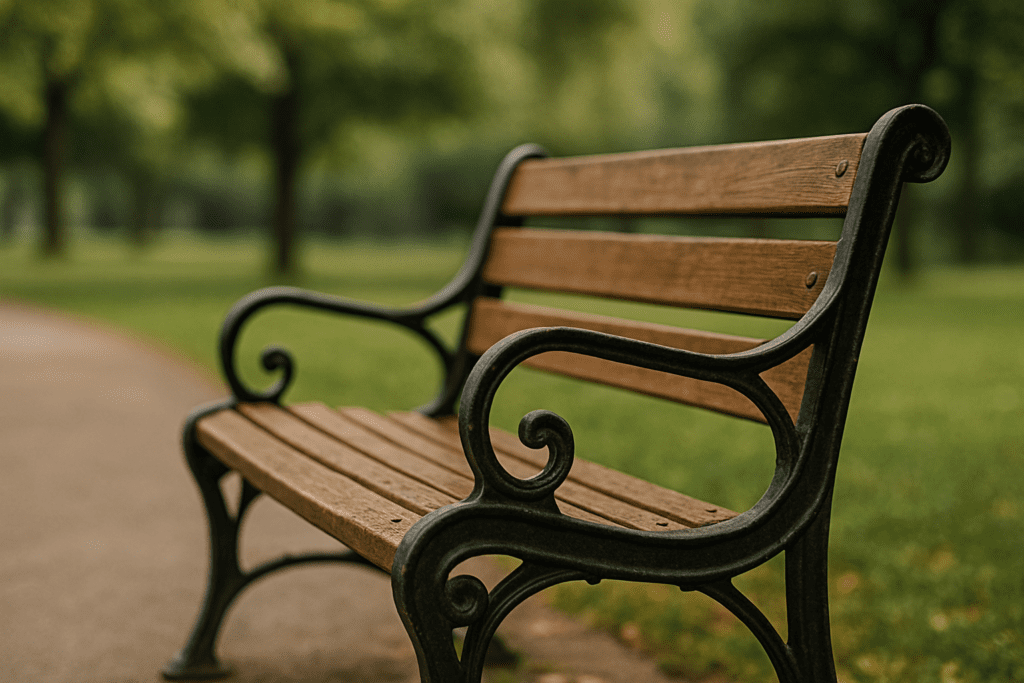 Wooden park bench with black iron arms beside a path, surrounded by soft-focus greenery on a cloudy day