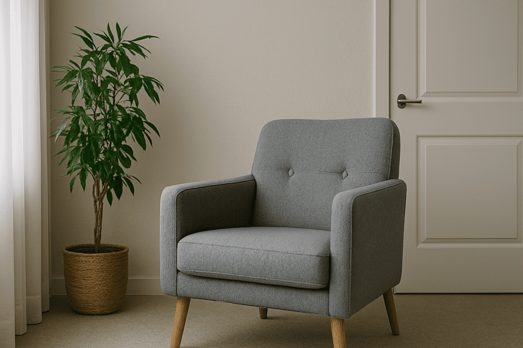 A minimalist therapy room with a gray armchair and a tall potted plant, lit by filtered daylight through sheer curtains and soft, neutral décor