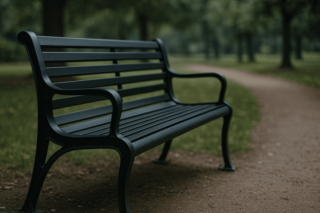 A black metal bench sits alone on a gravel path in a quiet park, surrounded by trees and dappled sunlight on a partly cloudy day