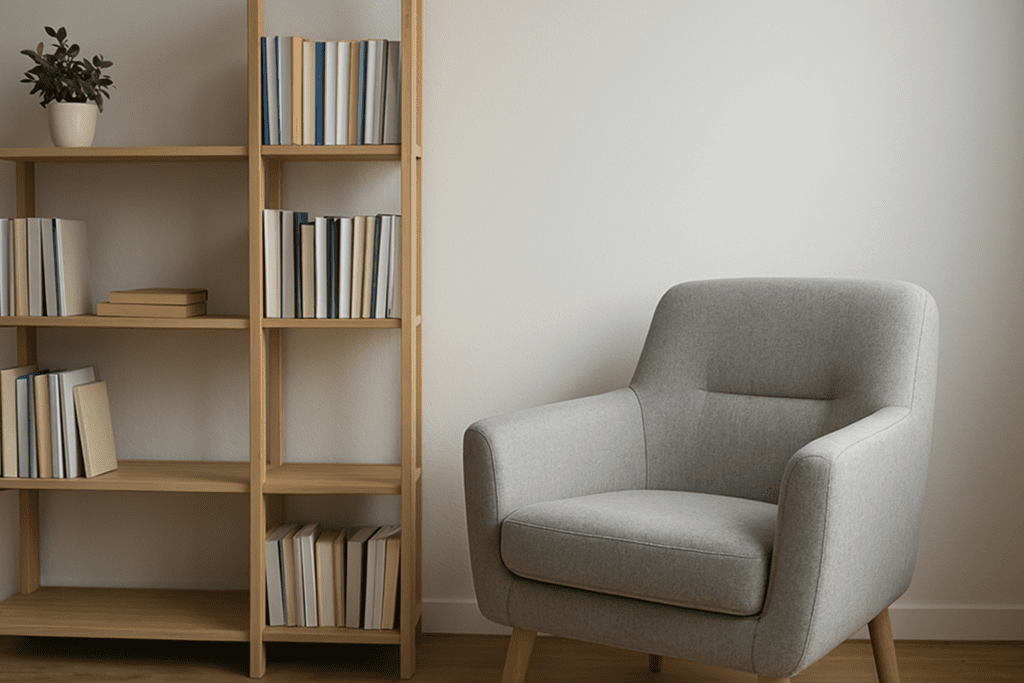 A gray armchair sits beside a modern bookshelf in a serene therapy space, enhanced by soft daylight and neutral tones